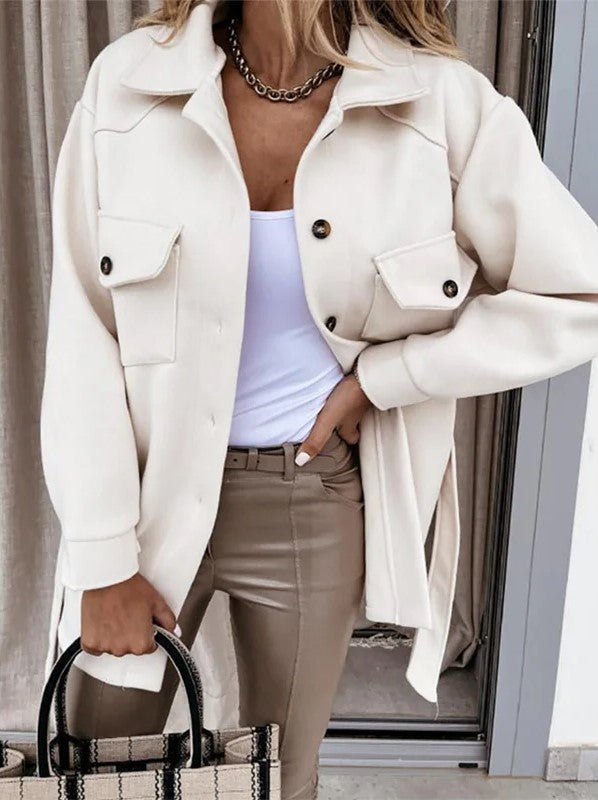 Femme portant une veste noire élégante, chemise blanche, pantalon beige, collier doré, tenant un sac à main à carreaux. Mode chic et moderne.